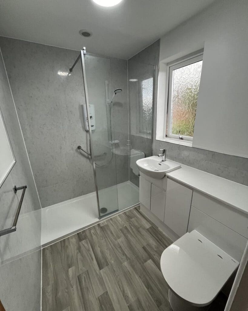 Modern bathroom with a walk-in shower, glass door, gray wall panels, white sink with cabinet, toilet, towel rail, and wood-look flooring. A frosted window lets in natural light.