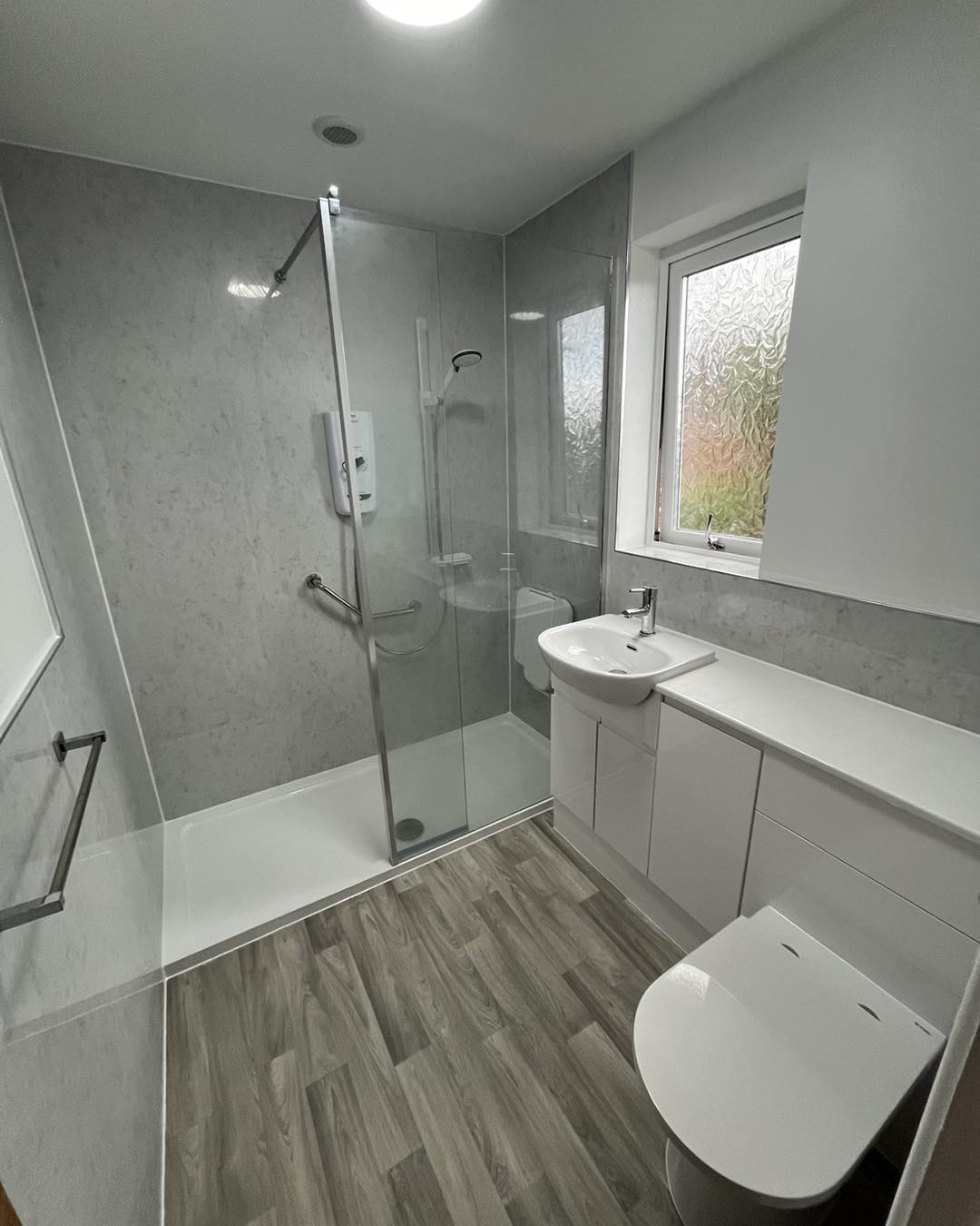 Modern bathroom with a walk-in shower, glass door, white sink, built-in cabinets, toilet, towel rail, and a window with frosted glass. The floor has a wood-look design.