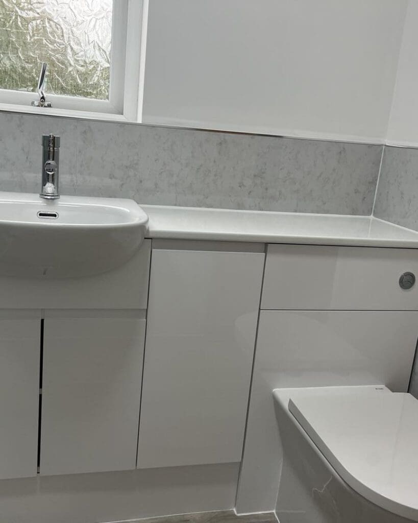 Modern bathroom with white cabinets, a sink with a chrome faucet, a toilet, and a frosted window above. The design is clean, minimal, and bright with light grey marble-effect wall panels.