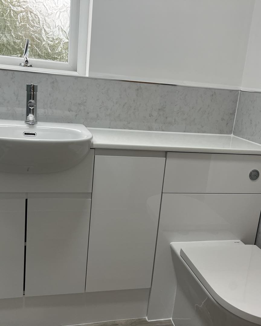 Modern bathroom with white cabinets, a white sink with chrome tap, and a white toilet. A frosted window is above the sink, allowing natural light into the room. Surfaces are clean and minimalistic.