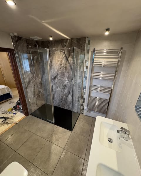 Modern bathroom with a glass shower enclosure, dark tiled floor, double sink, and a chrome towel radiator on the wall. Tools and materials are visible in the adjacent room, suggesting ongoing renovation.