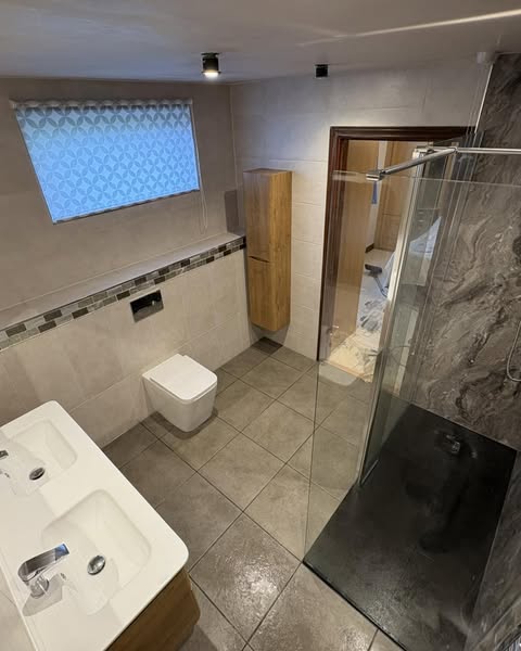 Modern bathroom with tiled floor, wall-mounted toilet, double sink, tall wooden cabinet, walk-in shower with glass door, and frosted window. Towels and materials are visible outside the bathroom door.