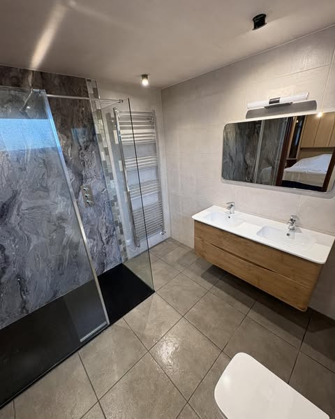 Modern bathroom with a glass shower, gray marble accent wall, heated towel rack, double sink vanity with mirror, and beige tile flooring.