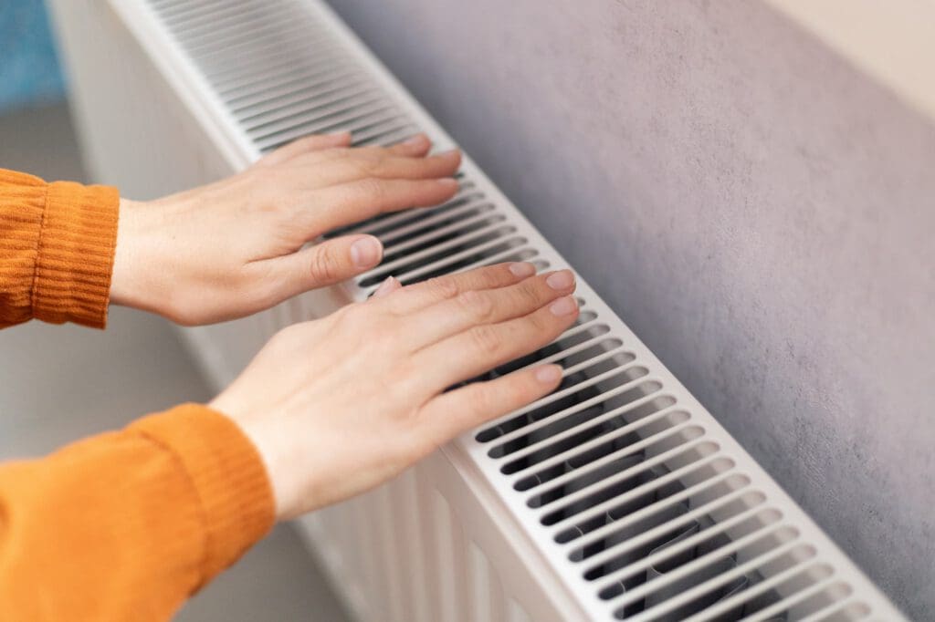 Two hands in an orange sweater are held above a white radiator, warming up indoors.