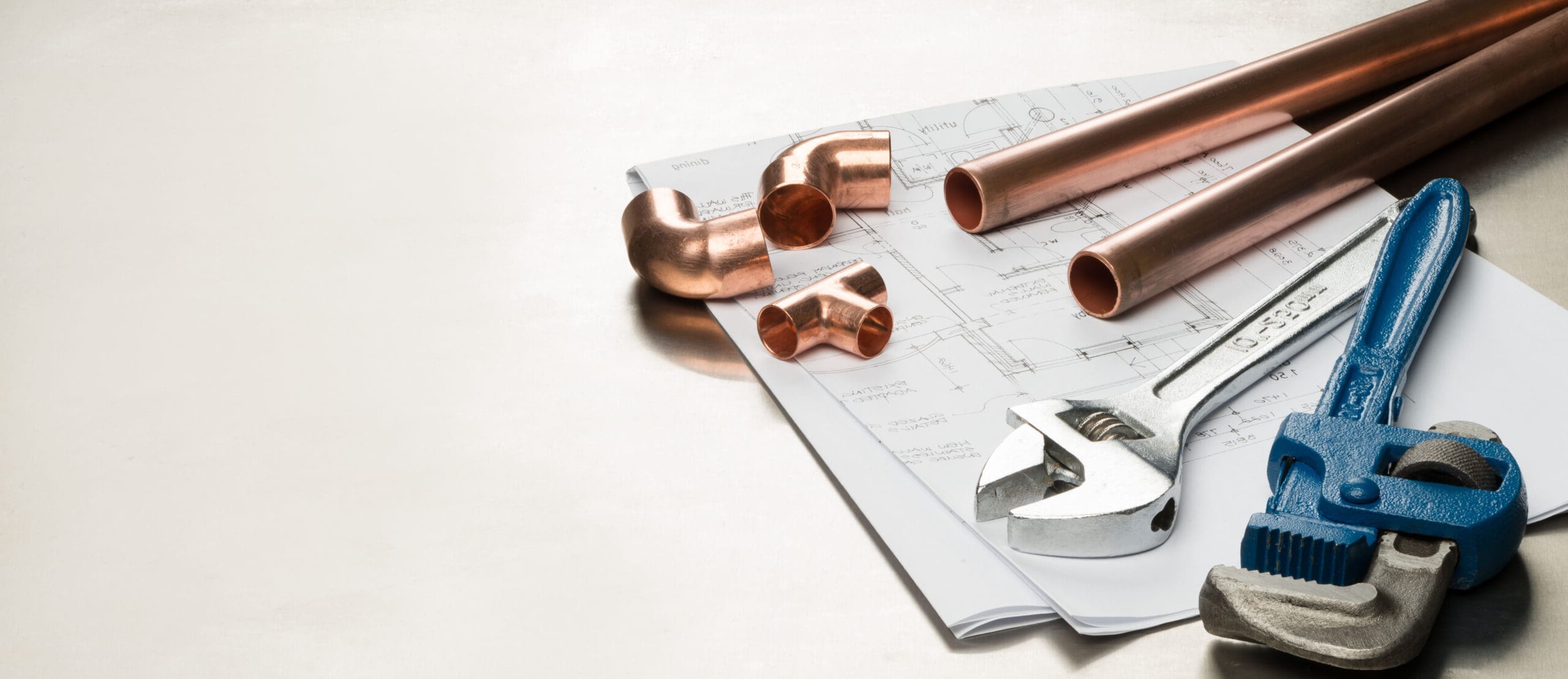 Copper pipes, pipe fittings, an adjustable wrench, and a pipe wrench are placed on top of plumbing blueprints on a light surface.