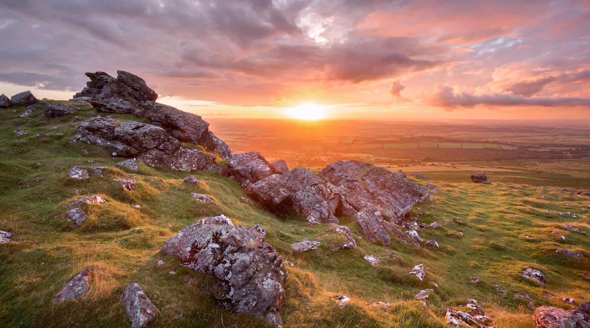 Rocky hillside with green grass at sunset, with the sun setting over a distant landscape of fields under a partly cloudy sky, casting warm orange and pink light across the scene.