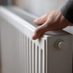 A person wearing a dark sweater touches the top of a white radiator attached to a wall, checking its temperature.