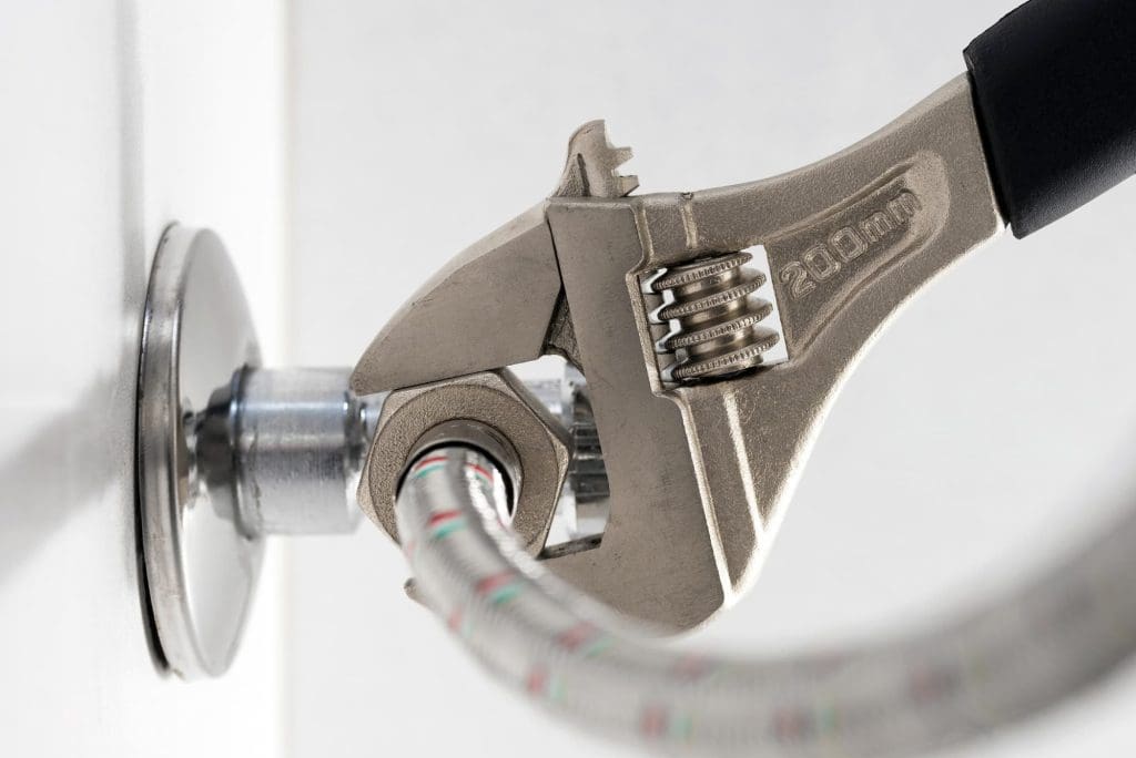 An adjustable wrench is tightening a metal nut on a flexible braided pipe connected to a wall fitting.