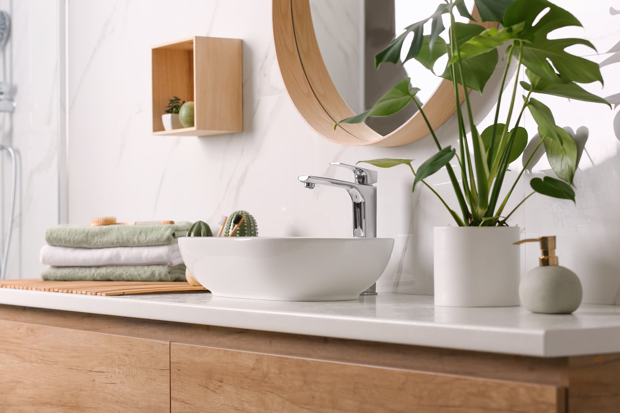 Modern bathroom with a white sink, chrome faucet, potted plant, soap dispenser, stacked towels, and a round mirror on a marble wall. Wooden cabinets and shelves add warmth to the decor.