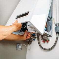A person adjusts or repairs a wall-mounted water heater or boiler, with the front panel open to reveal pipes, dials, and internal components.