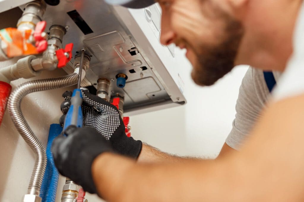 A plumber wearing gloves uses a wrench to repair or install pipes connected to a wall-mounted appliance, possibly a boiler or water heater. The plumber is focused on the task, and various pipes and valves are visible.