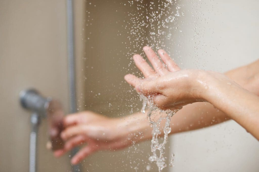 Close-up of a person’s hands feeling water from a shower, with water droplets visible in the air and a showerhead blurred in the background.