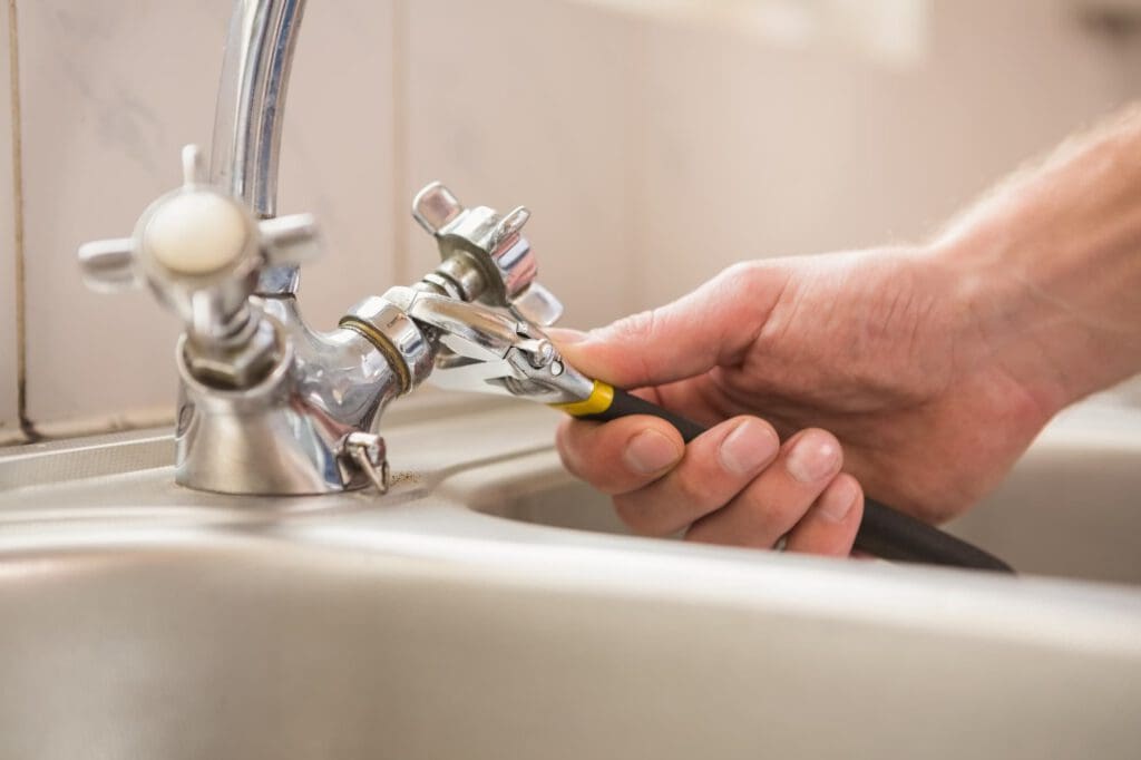 A close-up of a person's hand using a wrench to tighten a pipe fitting on a chrome kitchen faucet above a sink.