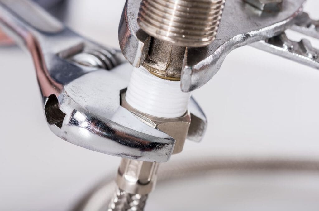 Close-up of two wrenches tightening a threaded plumbing fitting wrapped with white Teflon tape, attached to a braided metal hose.