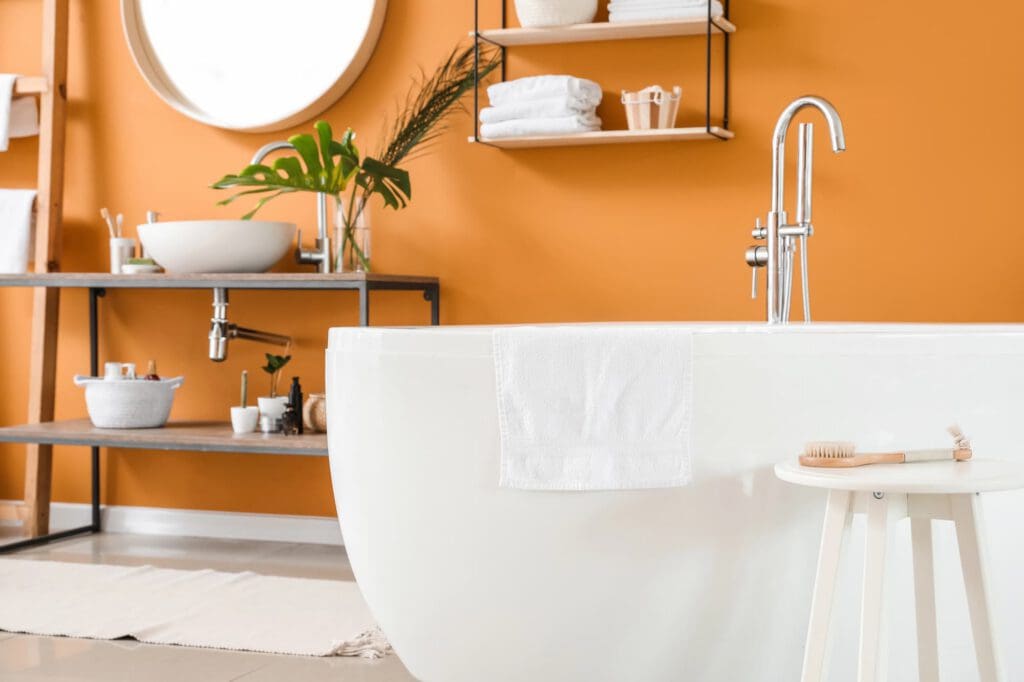 Modern bathroom with an orange wall, white bathtub, and towel. A small stool holds a brush. Shelves above the sink display towels, plants, and toiletries, creating a bright, organized, and cozy atmosphere.