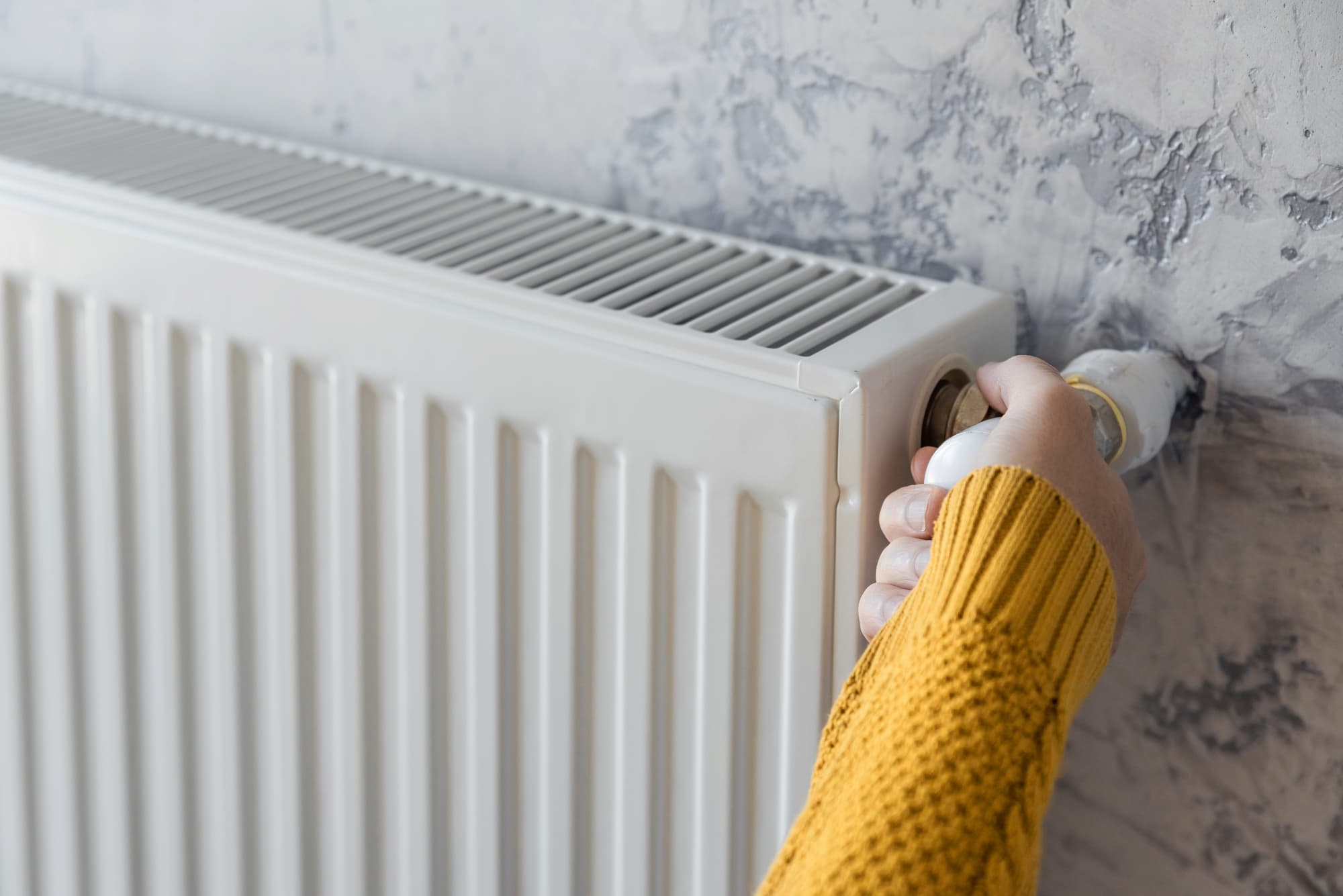 A person in a yellow sweater adjusts the valve on a white radiator attached to a textured grey wall.