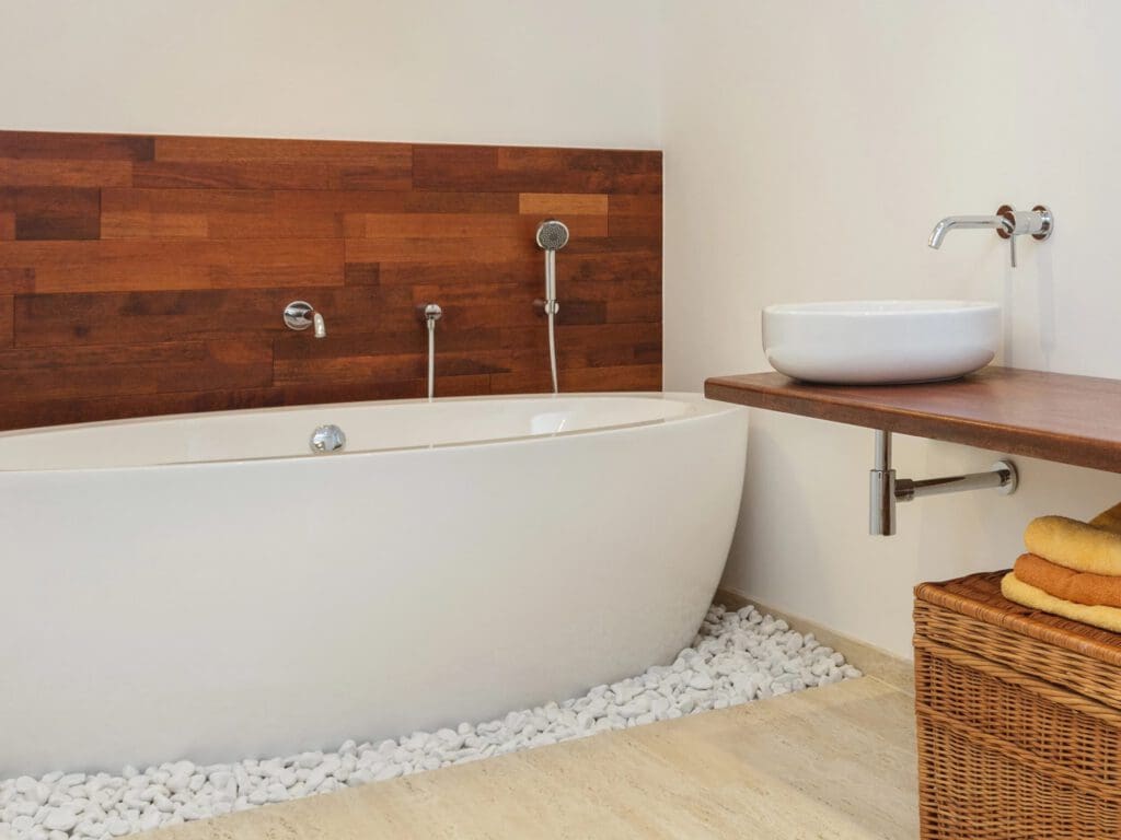 Modern bathroom with a white freestanding tub on white pebbles, wood accent wall, vessel sink on a wooden counter, and a wicker basket with folded yellow towels underneath.