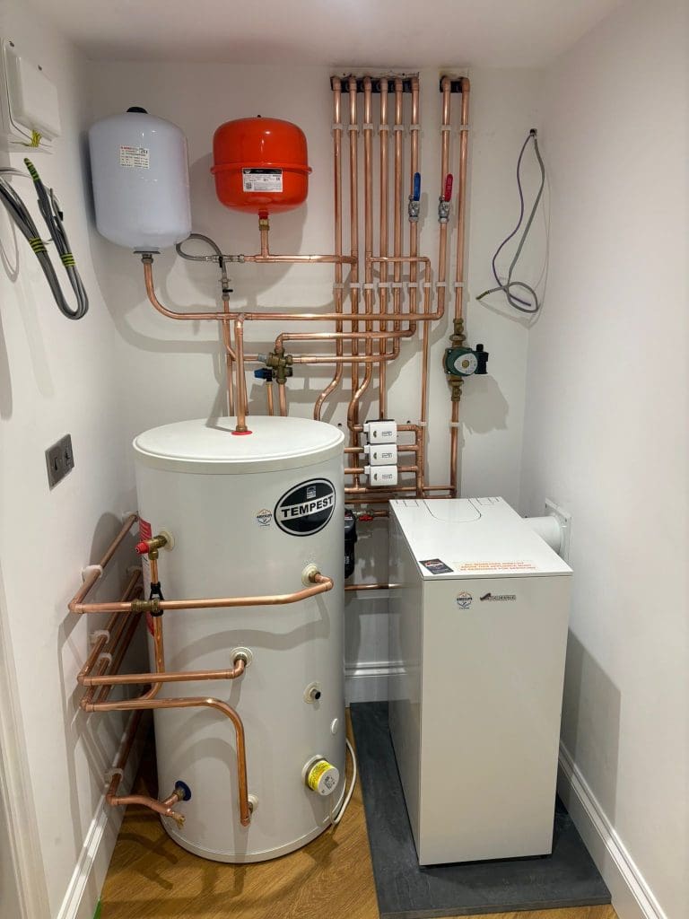 A neatly installed boiler and hot water cylinder system with copper pipes, a white Tempest tank, an expansion vessel, and control boxes in a white utility room with wooden flooring.