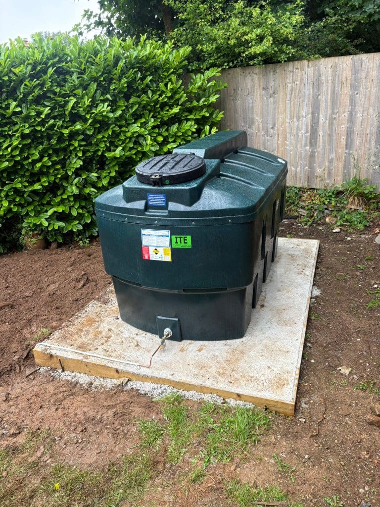 A large green plastic oil tank sits on a concrete slab in a garden, near a wooden fence and green bushes, with a black lid on top and a pipe extending from the tank.