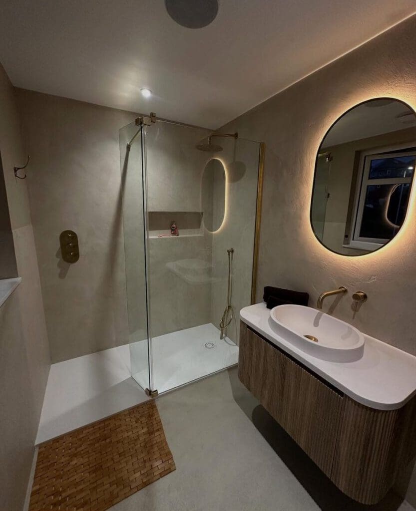 Modern bathroom with glass shower, oval mirror with soft backlighting, wall-mounted sink with gold fixtures, and a woven mat on the floor. A window is on the right wall, and the lighting is warm.