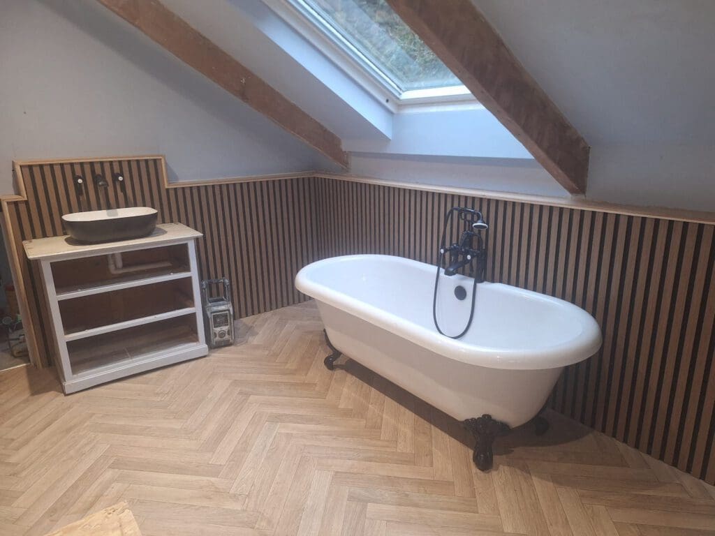 Modern attic bathroom with a freestanding white bathtub, angled skylight, wooden slat wall paneling, and a sink on a wooden stand. The floor has a light herringbone pattern.
