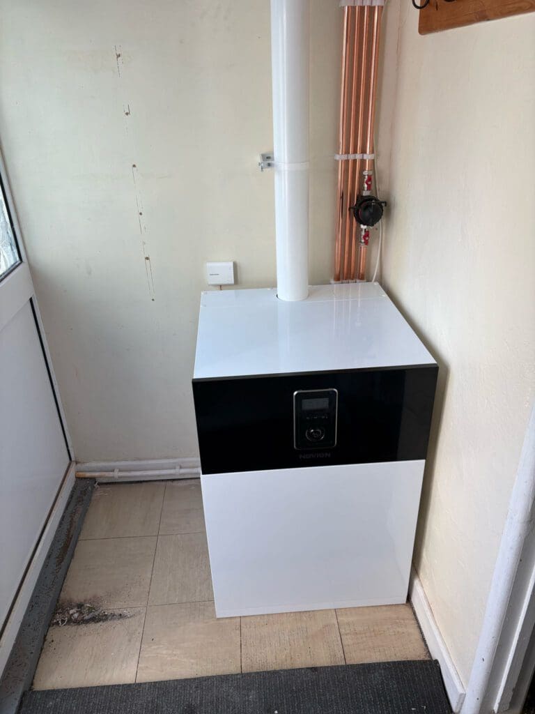 A modern boiler unit with a digital display is installed in the corner of a room. Copper pipes run vertically next to it, and a white vent pipe extends upward from the unit. The floor has beige tiles and a grey mat.