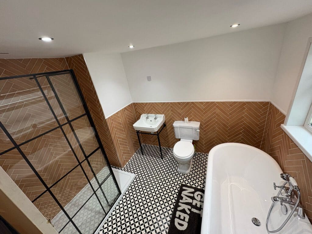 Modern bathroom with a glass shower, vintage sink, white toilet, and freestanding tub. Brown herringbone tiles, black and white patterned floor, and a rug saying “GET NAKED.”.