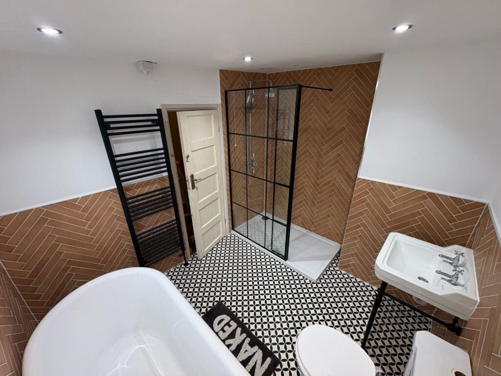 Modern bathroom with a freestanding bathtub, walk-in shower with glass doors, black towel rack, white sink, and a “NAKED” mat on a black and white patterned floor, and brown herringbone wall tiles.