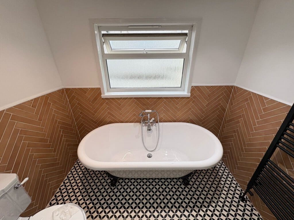 A white freestanding bathtub is centered against a brown herringbone-tiled wall, beneath a frosted window. The floor features a black and white geometric pattern.