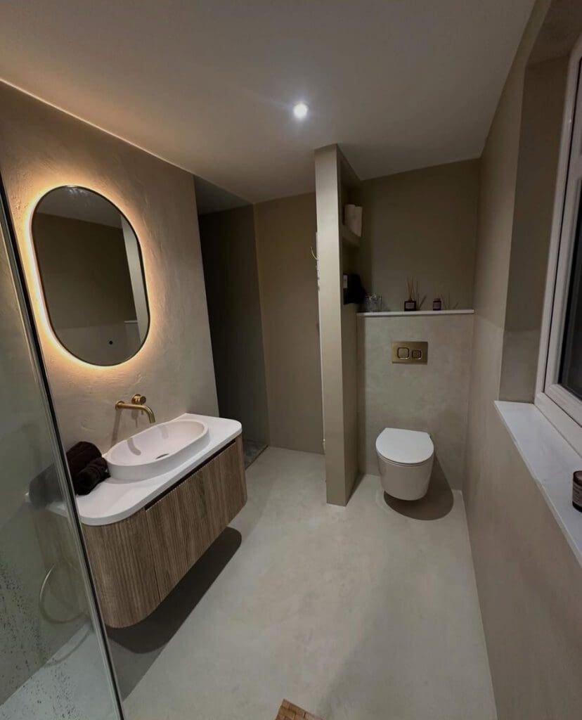 Modern bathroom with beige walls, a floating wooden vanity with a round mirror above, wall-mounted faucet, towel, wall-mounted toilet, and a window on the right. Minimalist design with soft, warm lighting.