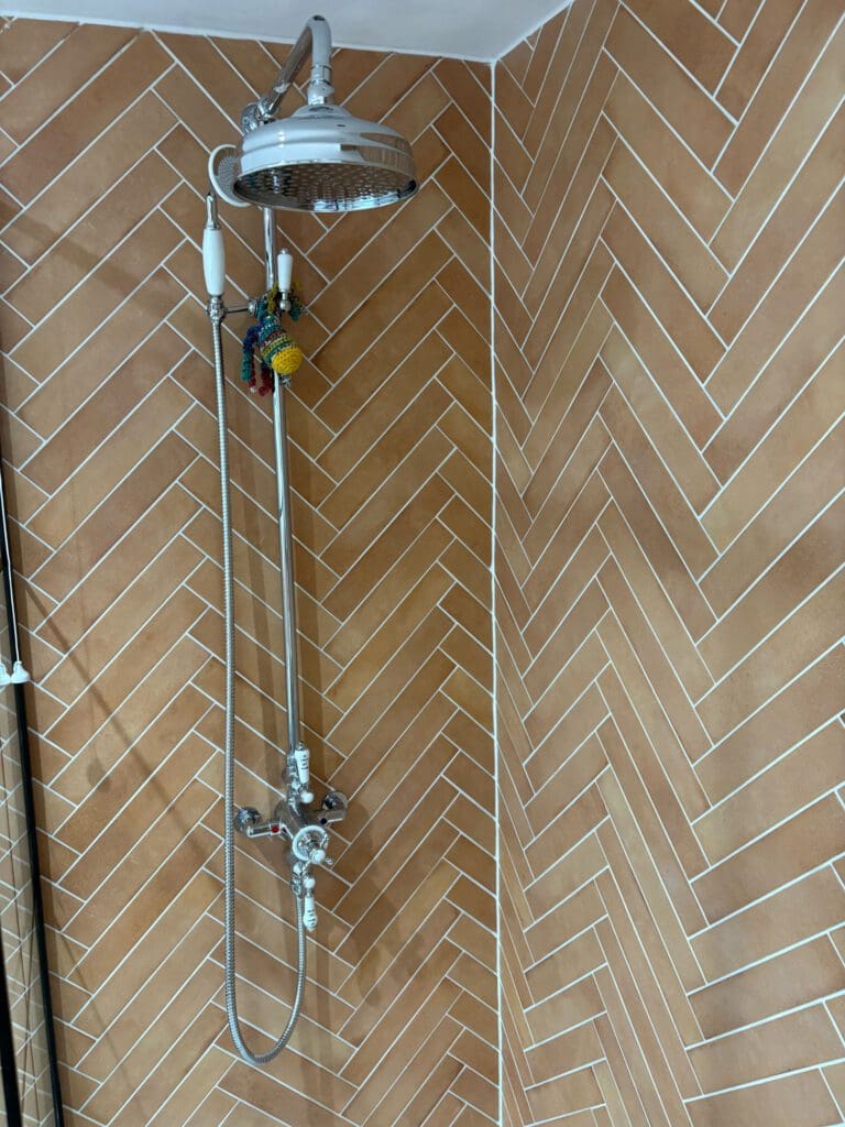 A shower with a round overhead showerhead and handheld attachment is mounted on a wall with brown herringbone tiles. Several colorful scrubbing sponges hang from the pipe.