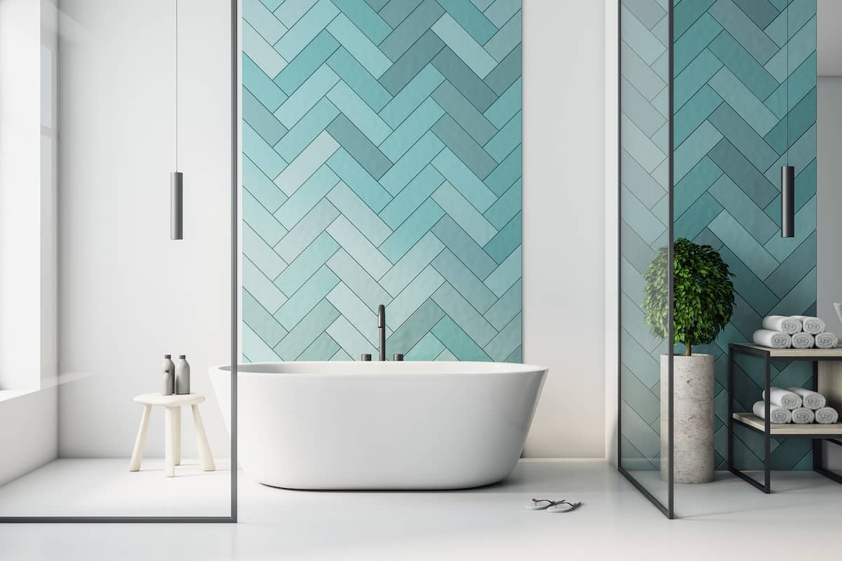 Modern bathroom with a white freestanding tub, turquoise herringbone tile accent wall, small stool, plant, glass dividers, and a shelf with neatly rolled towels. Natural light fills the clean, minimalistic space.